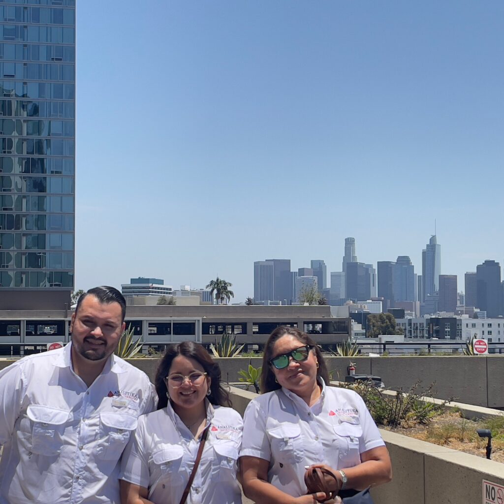 Three Analitika team members in white company shirts on a Koreatown rooftop, with the Downtown Los Angeles skyline behind them
