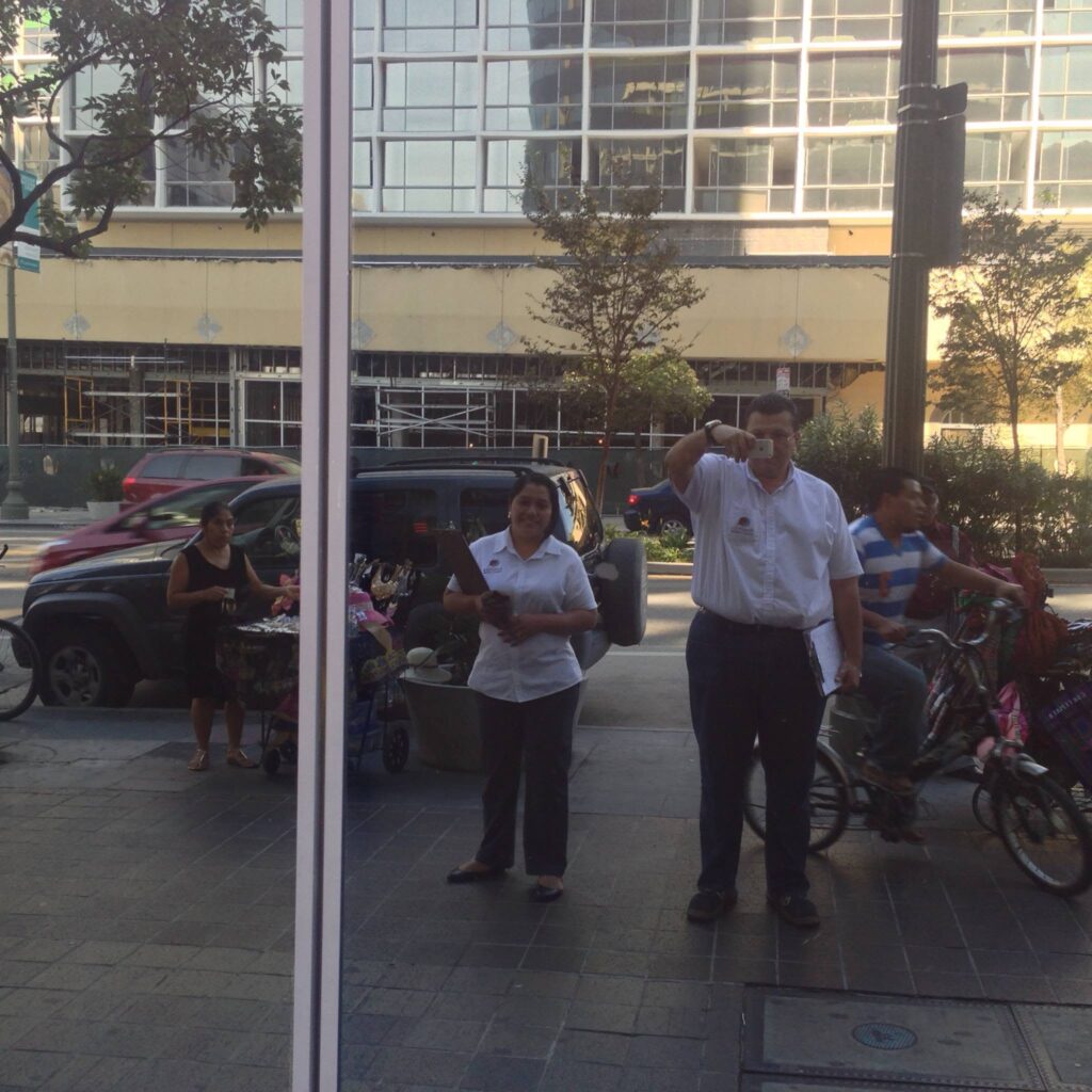 Storefront window reflection of two Analitika team members on a Los Angeles sidewalk, one of them taking the photo, with street vendors nearby