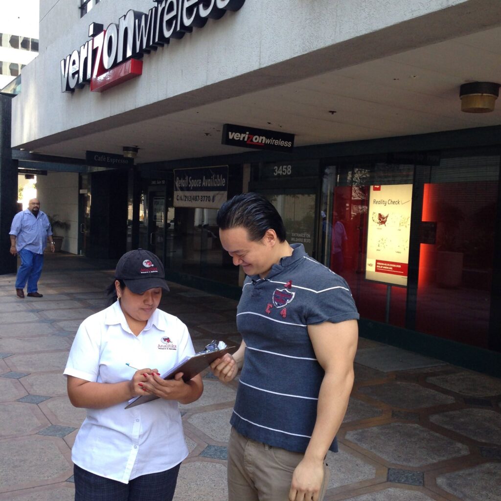 Analitika interviewer records answers on a clipboard while a respondent leans in to review the questionnaire on a Los Angeles sidewalk