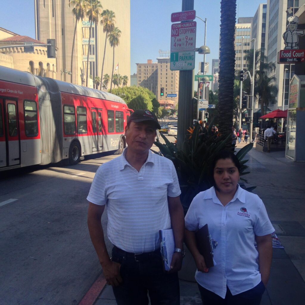 Two Analitika field interviewers with clipboards on Wilshire Boulevard as a Metro Rapid bus passes behind them, Los Angeles