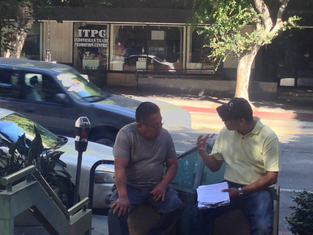 Analitika interviewer in a yellow polo holds a questionnaire while talking with a respondent seated beside him on a sidewalk bench, Wilshire Boulevard, Los Angeles